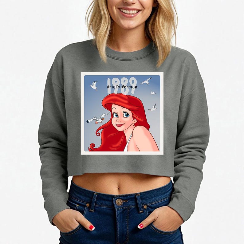 Pode incluir: Camisola cinzenta curta com um gr&aacute;fico de Ariel de A Pequena Sereia, com o texto "1989 Ariel's Version". O gr&aacute;fico mostra Ariel com cabelo ruivo e gaivotas contra um c&eacute;u azul. A camisola &eacute; de estilo casual.