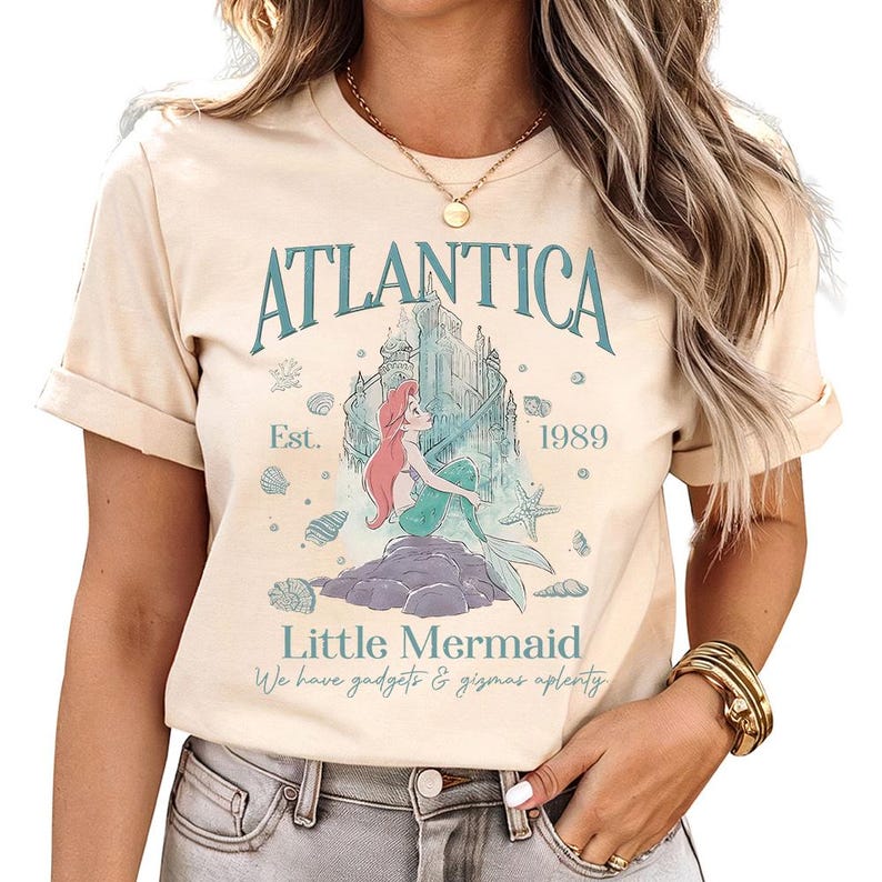 Puede incluir: Camiseta beige con un dise&ntilde;o gr&aacute;fico de una sirena de pelo rojo y cola verde, sentada en una roca. La camiseta tiene el texto "Atlantica", "Little Mermaid" y "We have gadgets & gizmos aplenty". El dise&ntilde;o incluye un castillo y conchas.