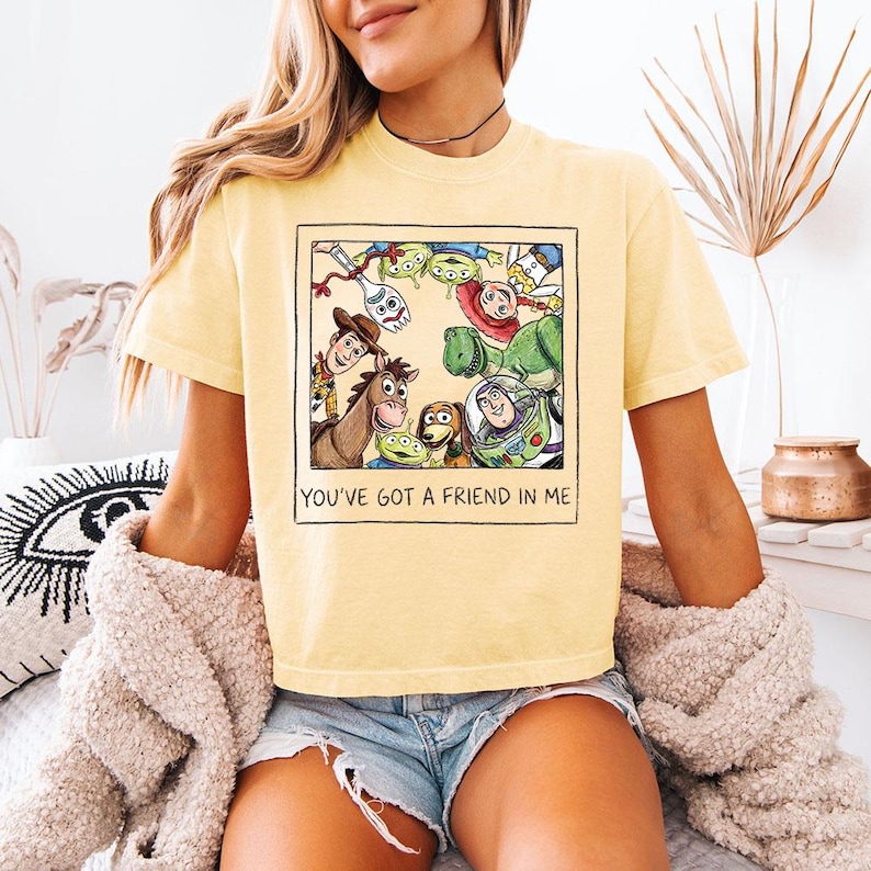 May include: Pale yellow t-shirt with a framed graphic of Toy Story characters, including Woody and Buzz Lightyear. The text "YOU'VE GOT A FRIEND IN ME" is printed below the graphic. The shirt has short sleeves.