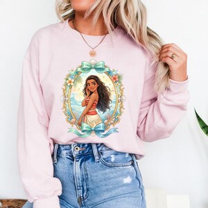 May include: A light pink sweatshirt featuring a framed illustration of a cartoon woman with long dark hair, wearing a red and white top and a white skirt. The frame has a teal bow at the top and bottom, with flowers and ocean waves.