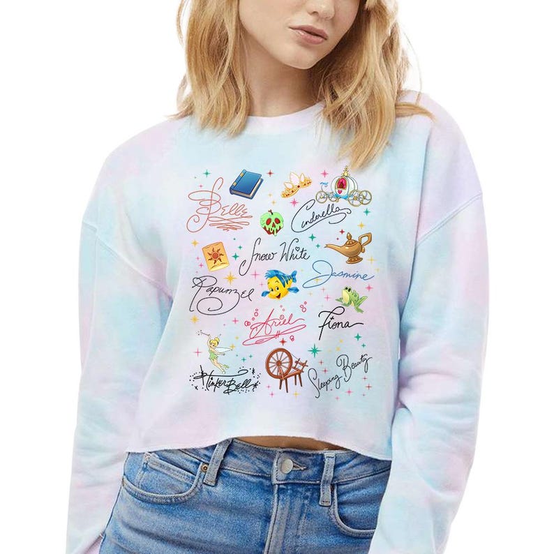 May include: A pastel tie-dye long-sleeve crop sweatshirt featuring a graphic print of various fairy tale character names in cursive script, including Belle, Cinderella, Snow White, Jasmine, Rapunzel, Ariel, Fiona, Tinker Bell, and Sleeping Beauty.