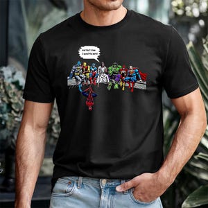 May include: Black t-shirt featuring a comic-style illustration of superheroes, including Batman, Superman, and Spider-Man. The image includes the text "And that's how I saved the world" in a speech bubble.