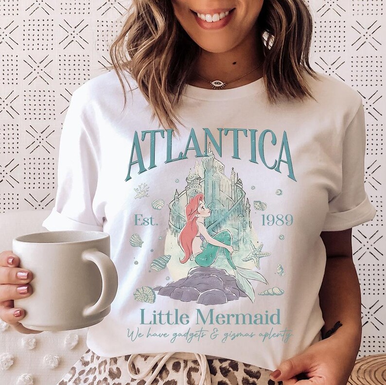 Puede incluir: Camiseta blanca con un gr&aacute;fico turquesa y azul claro de una sirena, un castillo y el texto "Atlantica" y "Little Mermaid". La camiseta tambi&eacute;n incluye el texto "Est. 1989" y "We have gadgets & gizmos aplenty".
