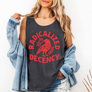 Könnte beinhalten: Dunkelgraues Tanktop mit dem roten Text "RADICALIZED BY BASIC DECENCY", der einen roten Vogel mit einem Zweig umkreist. Das Tanktop wird mit einer Jeansjacke getragen.