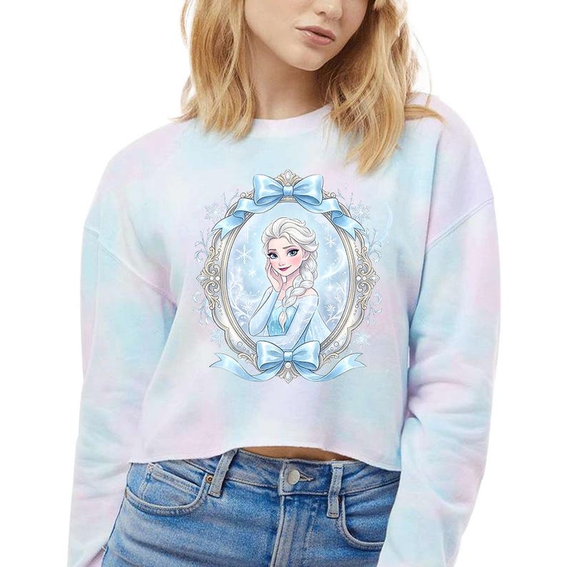 Op de afbeelding: Een cropped tie-dye sweatshirt in pastelblauw, roze en wit. De voorkant toont een ingelijste illustratie van een personage met wit haar en een blauwe jurk, versierd met blauwe strikken. Het ontwerp doet denken aan een populaire animatiefilm.