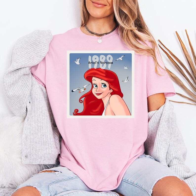 May include: Pink t-shirt featuring a square graphic of Ariel from The Little Mermaid, with red hair and blue eyes, surrounded by seagulls. The graphic includes the text "1989 Ariel's Version".