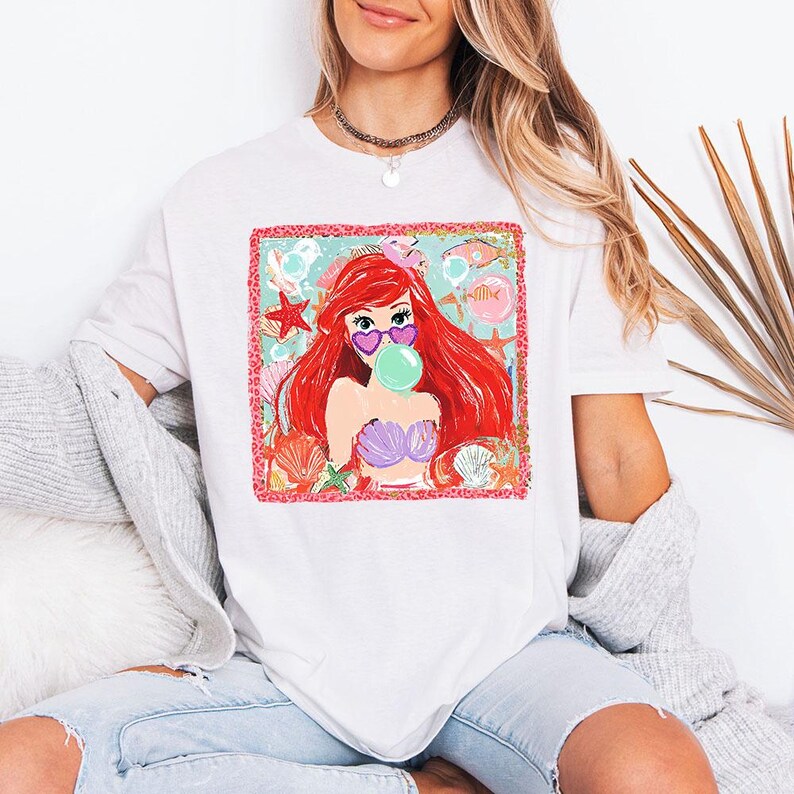 May include: White t-shirt featuring a colorful graphic of a mermaid with red hair, heart-shaped sunglasses, and a bubble gum bubble. The mermaid is wearing a purple seashell bra. The design includes starfish, seashells, and fish.