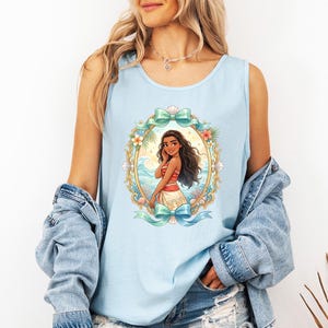 K&ouml;nnte beinhalten: Hellblaues Tanktop mit einer gerahmten Illustration einer Zeichentrickfigur mit langen dunklen Haaren, die ein rot-wei&szlig;es Oberteil und einen wei&szlig;en Rock tr&auml;gt. Der Rahmen enth&auml;lt einen goldenen Rand, Blumen und Meereswellen. Das Tanktop hat oben und unten eine hellblaue Schleife.