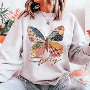 May include: A light gray crewneck sweatshirt featuring a large butterfly graphic on the chest. The butterfly wings are composed of various colorful patchwork fabric patterns, including floral, plaid, and gingham prints. The word Dolly is written below.