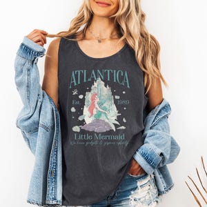 Op de afbeelding: Donkergrijze tanktop met de tekst "Atlantica" en "Little Mermaid" en een afbeelding van een zeemeermin en kasteel. De tanktop heeft ook de tekst "Est. 1989" en "We have gadgets & gizmos aplenty".