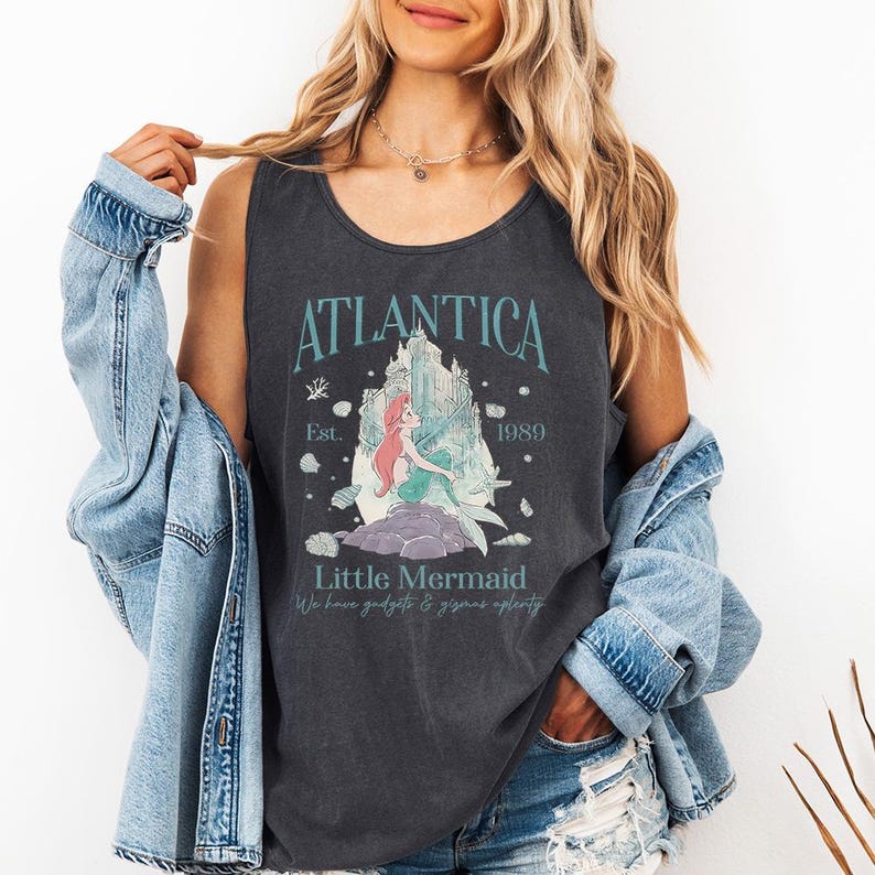 Op de afbeelding: Donkergrijze tanktop met de tekst "Atlantica" en "Little Mermaid" en een afbeelding van een zeemeermin en kasteel. De tanktop heeft ook de tekst "Est. 1989" en "We have gadgets & gizmos aplenty".
