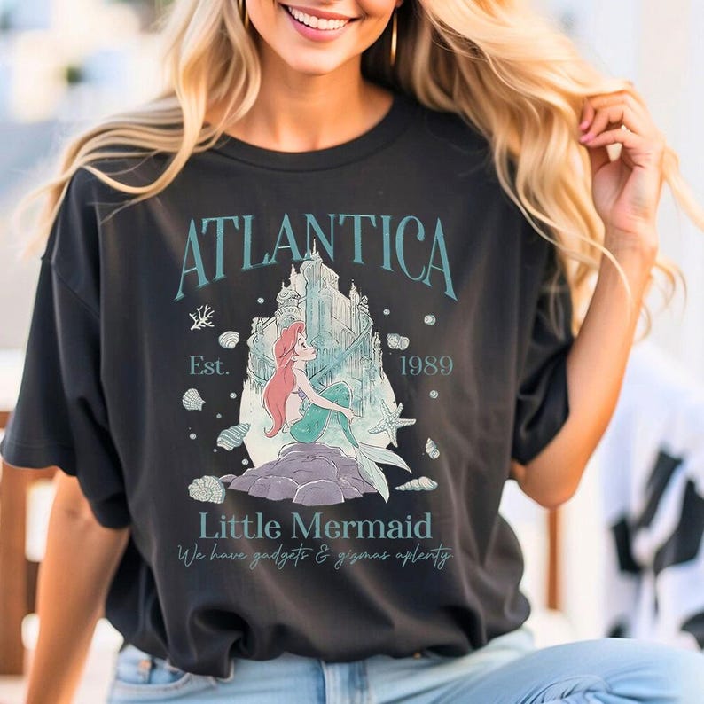Op de afbeelding: Zwart T-shirt met een afbeelding van de Kleine Zeemeermin, de tekst "Atlantica" en "Little Mermaid". Het ontwerp bevat een kasteel, schelpen en het jaar 1989. Het shirt heeft ook de zin "We have gadgets & gizmos aplenty."