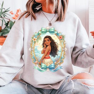 May include: A light gray sweatshirt featuring a framed illustration of a woman with long dark hair, wearing a red and white outfit. The illustration is surrounded by a gold frame with flowers, seashells, and blue bows.