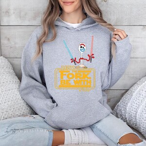 May include: A heather gray hooded sweatshirt featuring a graphic of a white plastic spork character with red pipe cleaner arms. The character holds a blue and a red lightsaber. Below, yellow text reads: MAY THE FORK BE WITH YOU.