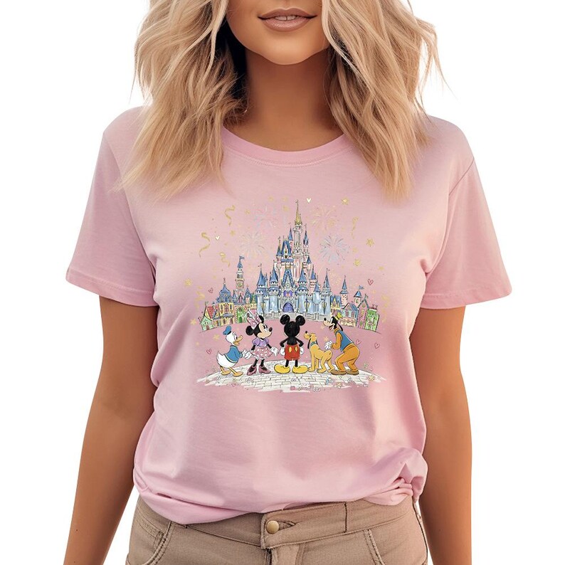 Op de afbeelding: Lichtroze T-shirt met een kleurrijke afbeelding van Disney-personages Mickey Mouse, Minnie Mouse, Donald Duck en Goofy voor een kasteel. Het ontwerp bevat vuurwerk en gebouwen.