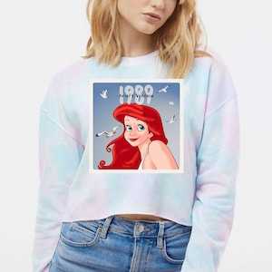 Pode incluir: Um moletom cropped tie-dye com um gr&aacute;fico de Ariel de A Pequena Sereia. O gr&aacute;fico apresenta Ariel com cabelo ruivo, olhos azuis e o texto "1989 Ariel's Version" com gaivotas no fundo.