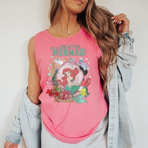 May include: A bright pink sleeveless tank top featuring a graphic print of The Little Mermaid characters including Ariel in a shell, King Triton, Ursula, Sebastian, and Flounder, with bubbles and coral accents printed on the front of the garment.