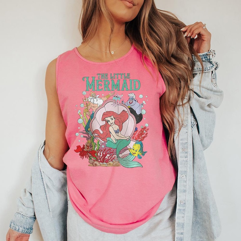 May include: A bright pink sleeveless tank top featuring a graphic print of The Little Mermaid characters including Ariel in a shell, King Triton, Ursula, Sebastian, and Flounder, with bubbles and coral accents printed on the front of the garment.