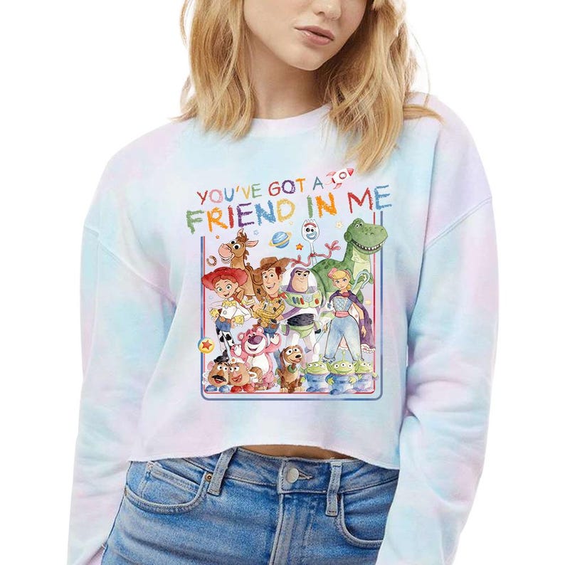 May include: A tie-dye cropped sweatshirt featuring a colorful graphic of characters from the animated film "Toy Story." The text "YOU'VE GOT A FRIEND IN ME" is displayed above the characters in a rainbow font.