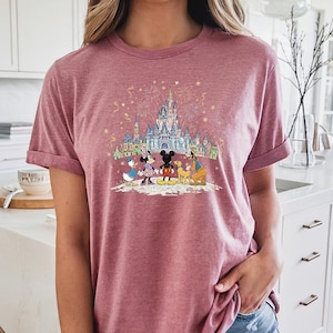 Op de afbeelding: Een bordeauxrood T-shirt met een kleurrijke afbeelding van Disney-personages voor een kasteel met vuurwerk. De afbeelding bevat Mickey en Minnie Mouse, Donald Duck, Goofy en Pluto. Het shirt heeft opgerolde mouwen.