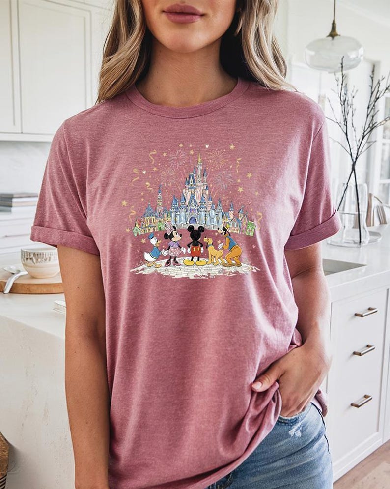 Op de afbeelding: Een bordeauxrood T-shirt met een kleurrijke afbeelding van Disney-personages voor een kasteel met vuurwerk. De afbeelding bevat Mickey en Minnie Mouse, Donald Duck, Goofy en Pluto. Het shirt heeft opgerolde mouwen.