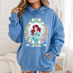 Puede incluir: Sudadera con capucha azul claro con una ilustraci&oacute;n enmarcada de una sirena con cabello rojo y un vestido verde azulado. El marco dorado tiene lazos verde azulado. La sudadera tiene una capucha con cord&oacute;n y un bolsillo delantero.