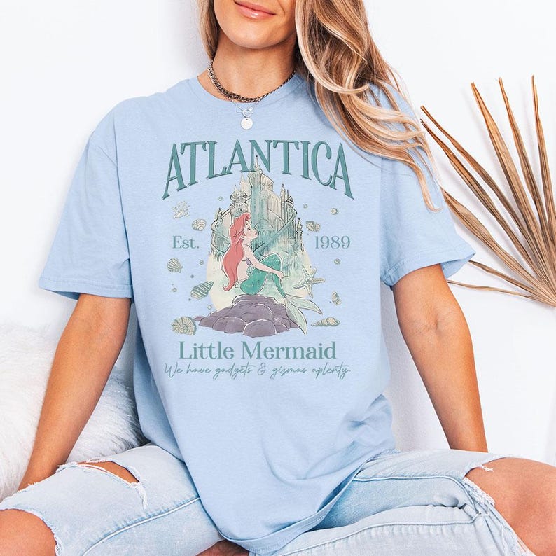 May include: Light blue t-shirt featuring a graphic of a mermaid with red hair and a green tail, sitting on a rock with a castle in the background. The shirt has the text "Atlantica" and "Little Mermaid" with the year "1989".