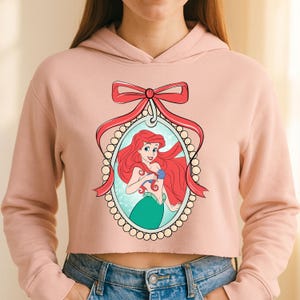 Puede incluir: Sudadera corta de color rosa claro con una ilustraci&oacute;n de dibujos animados de Ariel de La Sirenita. La ilustraci&oacute;n est&aacute; enmarcada por un lazo rojo y un &oacute;valo con perlas. Ariel tiene el pelo rojo y una cola verde.