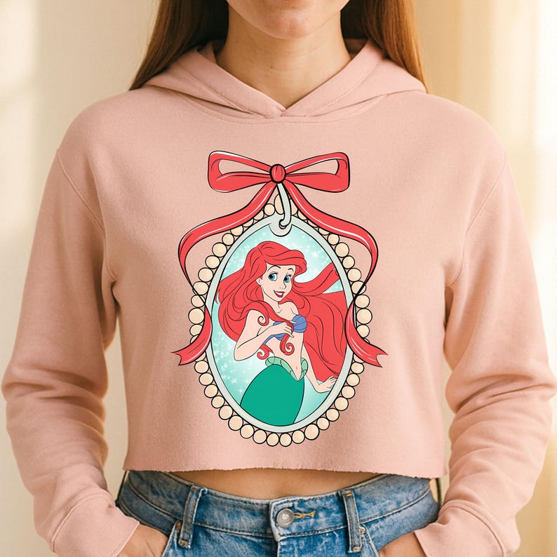 Puede incluir: Sudadera corta de color rosa claro con una ilustraci&oacute;n de dibujos animados de Ariel de La Sirenita. La ilustraci&oacute;n est&aacute; enmarcada por un lazo rojo y un &oacute;valo con perlas. Ariel tiene el pelo rojo y una cola verde.