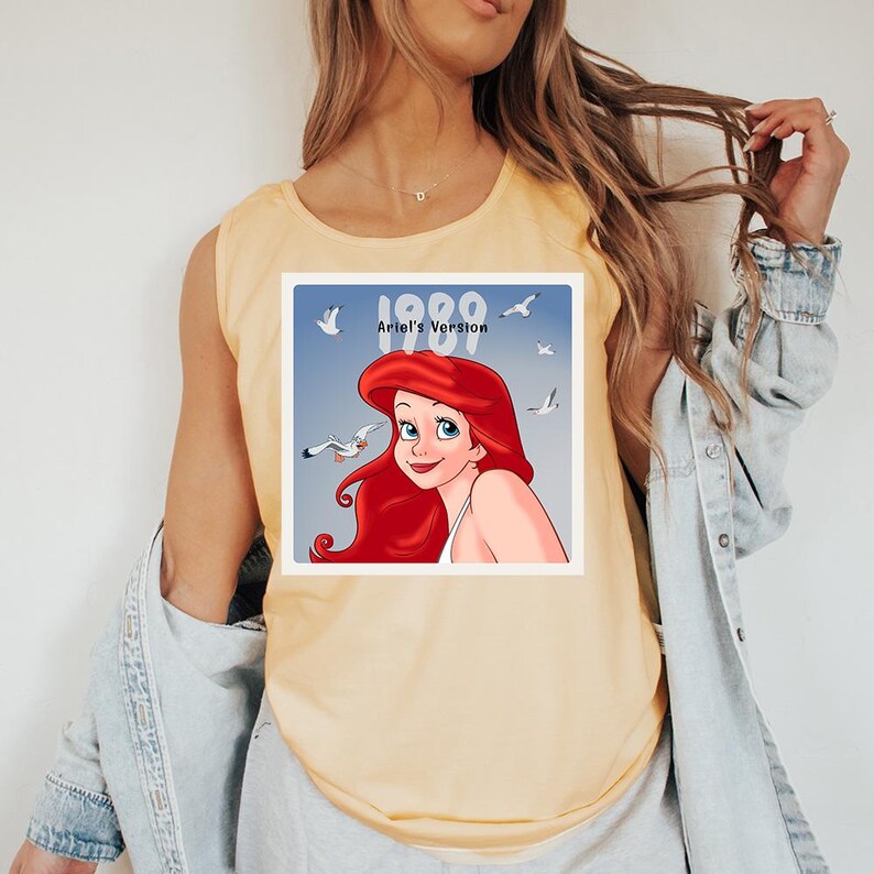 May include: Yellow tank top featuring a graphic of Ariel from The Little Mermaid, with the text "1989 Ariel's Version" and seagulls on a blue background. The shirt is a light, solid color.