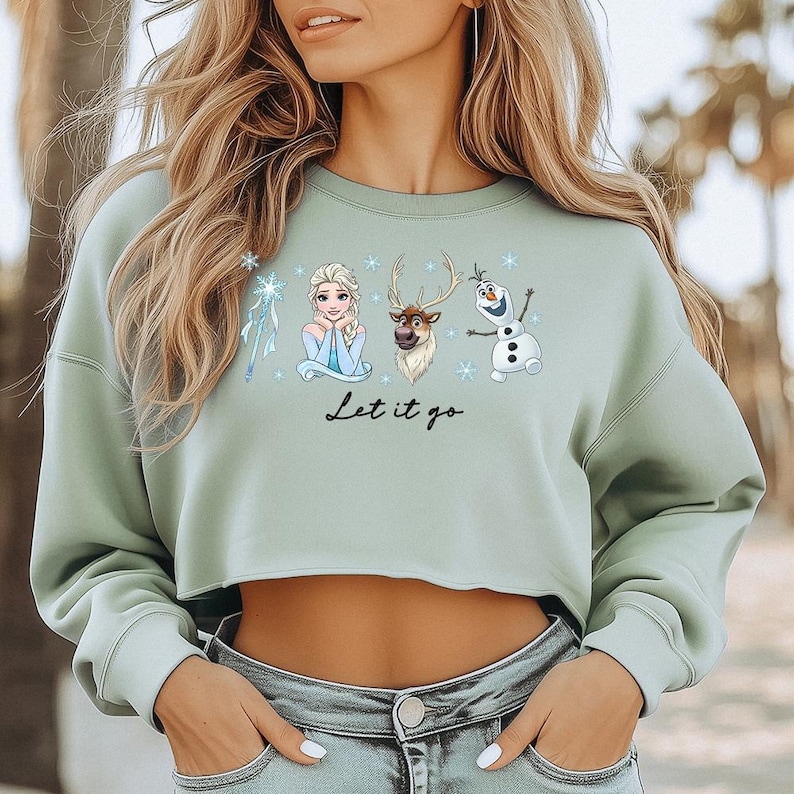 May include: Sage green cropped sweatshirt featuring Elsa, Sven, and Olaf from Frozen. The design includes snowflakes and the phrase "Let it go" in black script. The sweatshirt is a casual, comfortable style.