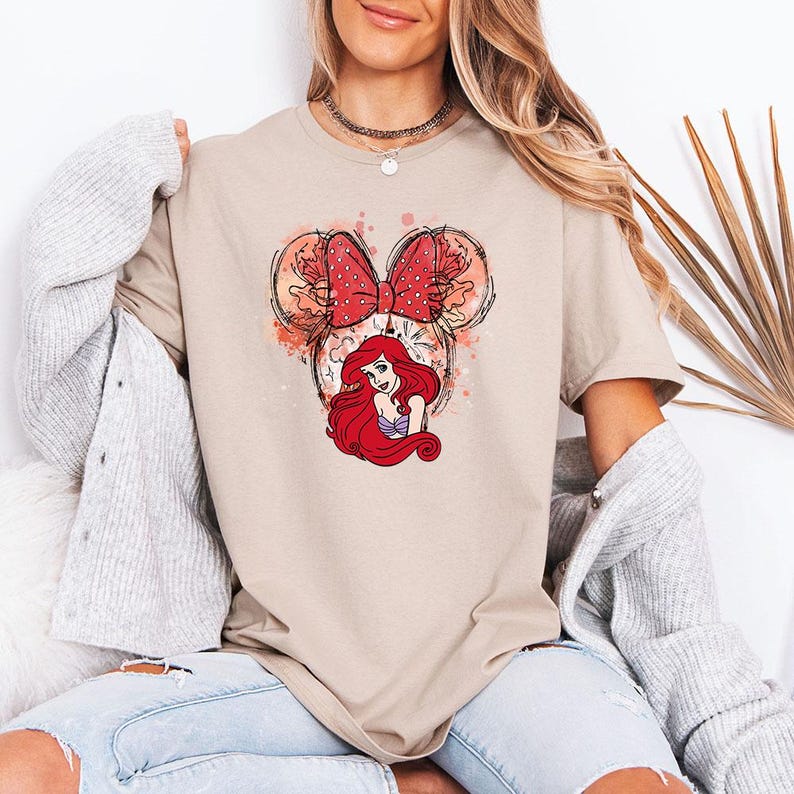 Puede incluir: Camiseta beige con un gr&aacute;fico de Ariel de La Sirenita. El dise&ntilde;o incluye un lazo rojo de lunares, orejas de Mickey Mouse y detalles de acuarela. La camiseta es una prenda informal.
