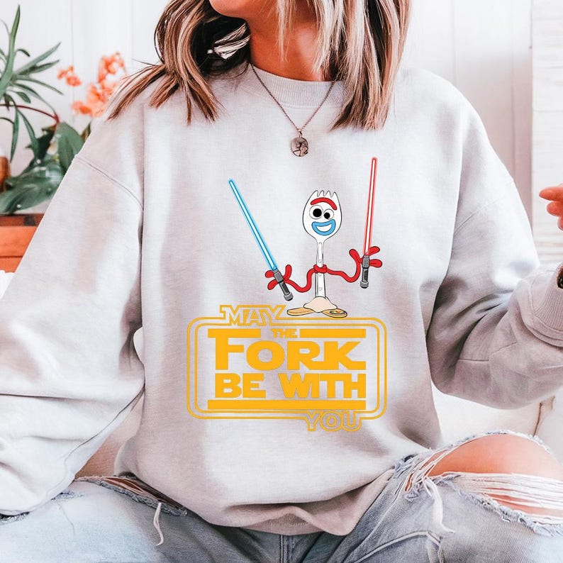 May include: A light grey crewneck sweatshirt featuring a graphic of the character Forky holding a blue lightsaber in one hand and a red lightsaber in the other. Below the character, the yellow text reads, "MAY THE FORK BE WITH YOU."