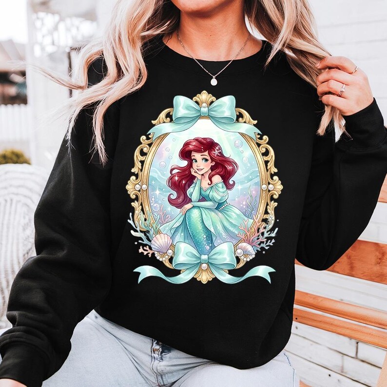 May include: Black sweatshirt featuring a framed illustration of a mermaid with red hair and a teal dress. The illustration is surrounded by a gold frame and teal bows. The mermaid is sitting in a sea scene with seashells and coral.