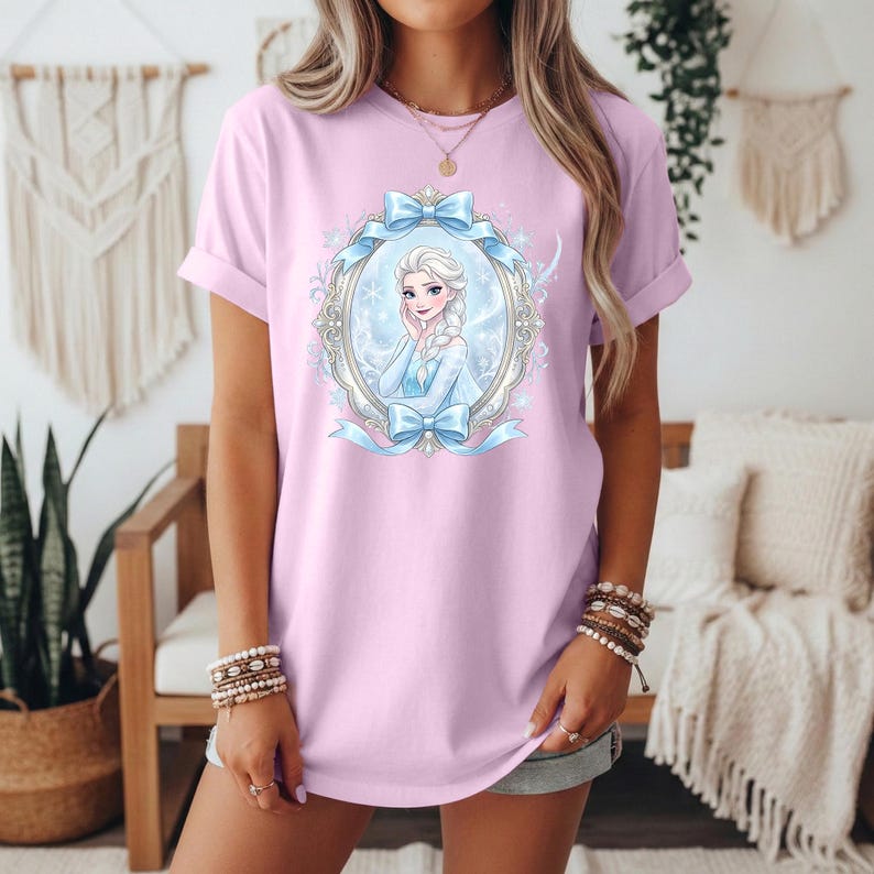 May include: A pale pink t-shirt with a cartoon image of a princess in an oval frame with blue bows and accents. The princess has blonde hair and wears a blue dress. The shirt has rolled-up sleeves.
