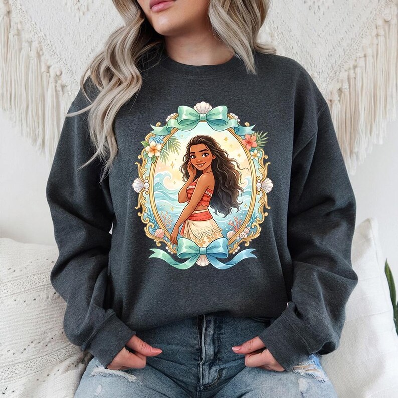 May include: Dark gray sweatshirt featuring a cartoon illustration of a woman in a red and white top and a cream skirt, framed by a gold oval with seashells, flowers, and a turquoise bow. The design is set against a backdrop of ocean waves and a sunset.