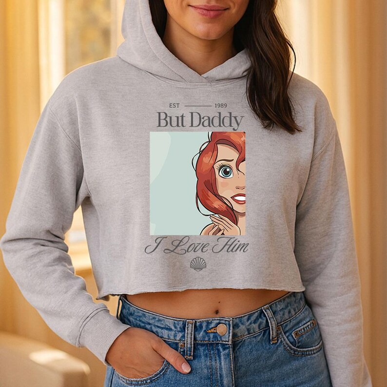 K&ouml;nnte beinhalten: Grauer Cropped-Hoodie mit dem Text "But Daddy" &uuml;ber einem Cartoon-Bild einer rothaarigen Figur. Unter dem Bild steht der Text "I Love Him". Der Hoodie hat eine Kapuze mit Kordelzug und lange &Auml;rmel.