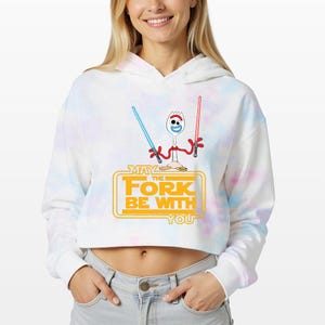 May include: A white and pastel tie-dye cropped hoodie featuring a graphic of a spork character holding a blue and a red lightsaber. Below the character, the yellow text reads MAY THE FORK BE WITH YOU, styled in a classic science fiction movie font.