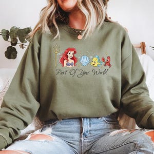 May include: An olive green sweatshirt featuring a graphic of Ariel, Flounder, Sebastian, and a seashell. The phrase "Part Of Your World" is printed below the characters. The sweatshirt is made of a soft material.