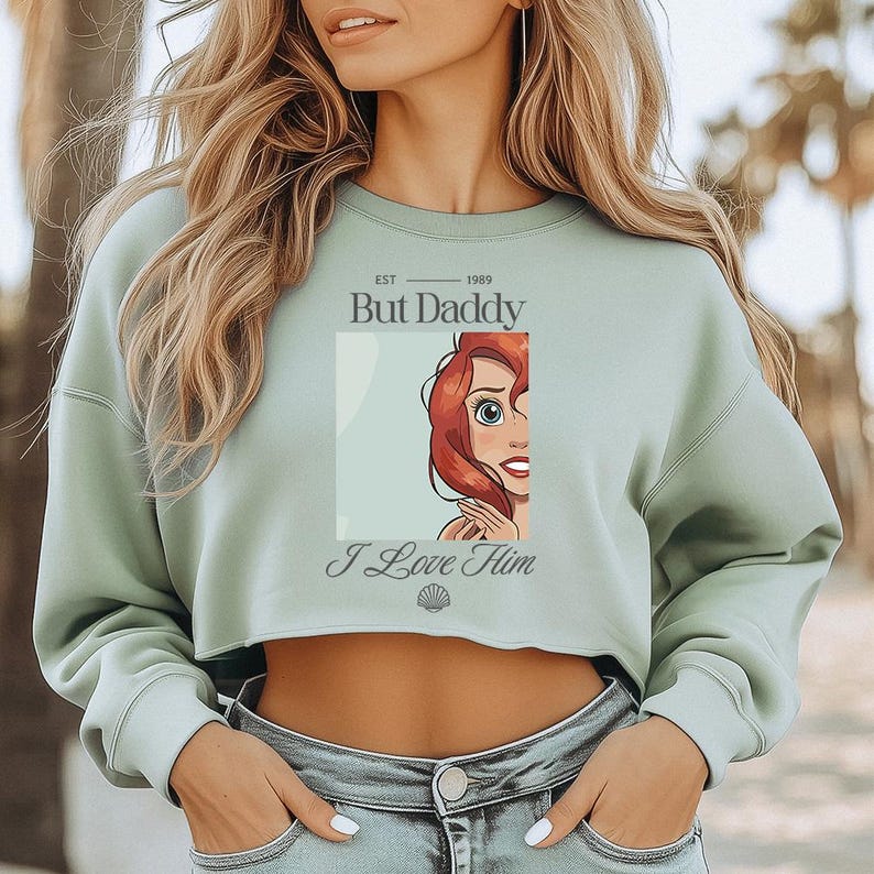 May include: A sage green cropped sweatshirt with the text "But Daddy" above a cartoon image of a red-haired character. Below the image is the text "I Love Him" and a seashell graphic. The sweatshirt has long sleeves.