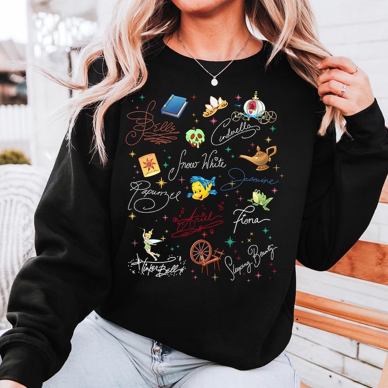May include: A black long-sleeve sweatshirt featuring a colorful graphic print of various fairy tale icons and character names written in cursive script, including Belle, Cinderella, Snow White, Jasmine, Rapunzel, Ariel, Fiona, and Tinker Bell.