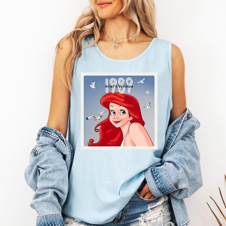May include: Light blue tank top featuring a graphic of Ariel from The Little Mermaid. The graphic includes the text "1989 Ariel's Version" with an illustration of Ariel and seagulls. The tank top is sleeveless.
