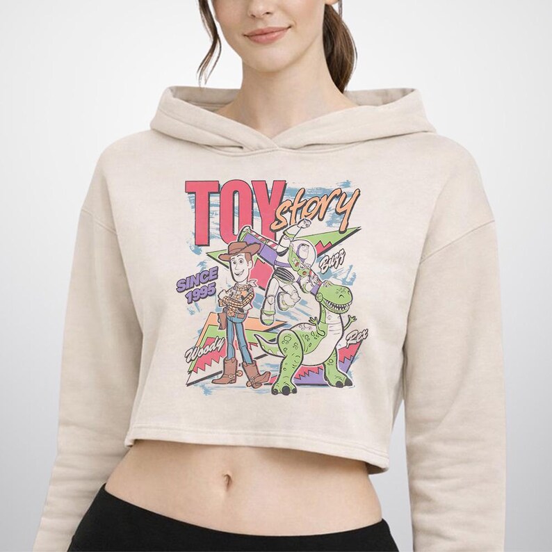 May include: A beige cropped hoodie featuring a graphic print of Toy Story characters Woody, Buzz Lightyear, and Rex. The design includes the text "TOY story", "SINCE 1995", "Woody", "Buzz", and "Rex" in colourful, retro-style lettering and geometric shapes.