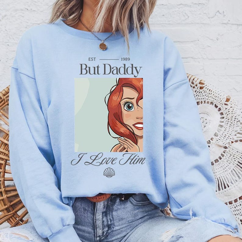 May include: Light blue sweatshirt with the text "But Daddy I Love Him" and a graphic of a red-haired cartoon character. The text "EST 1989" is above the main text. The graphic is in a rectangular frame.