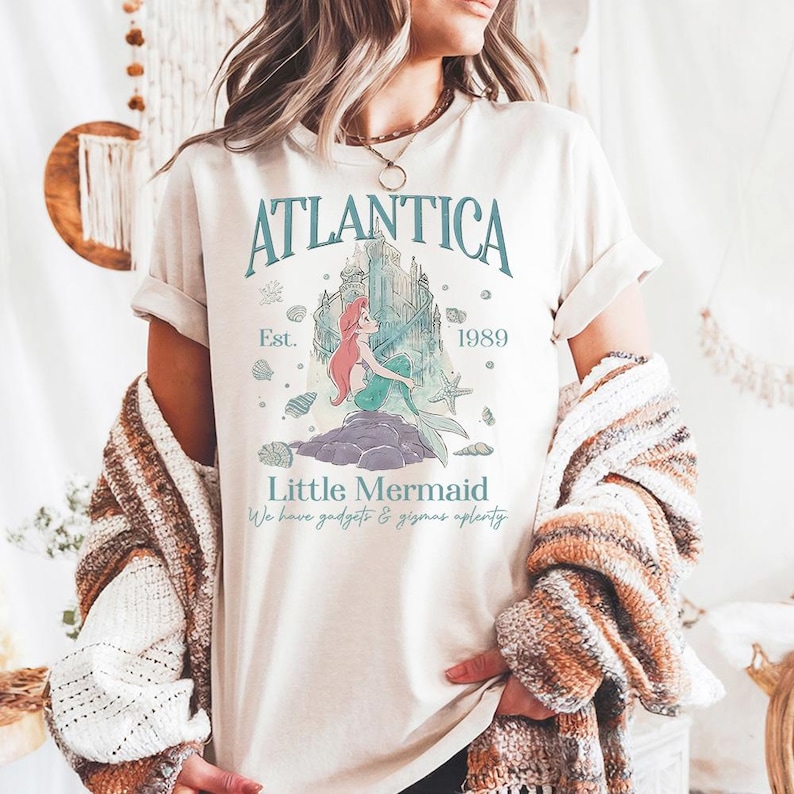 Op de afbeelding: Cr&egrave;mekleurig T-shirt met een teal-kleurige afbeelding met het woord "Atlantica" boven een illustratie van een zeemeermin en een kasteel. De tekst "Little Mermaid" en "We have gadgets & gizmos aplenty" zijn ook aanwezig. Het shirt heeft opgerolde mouwen.