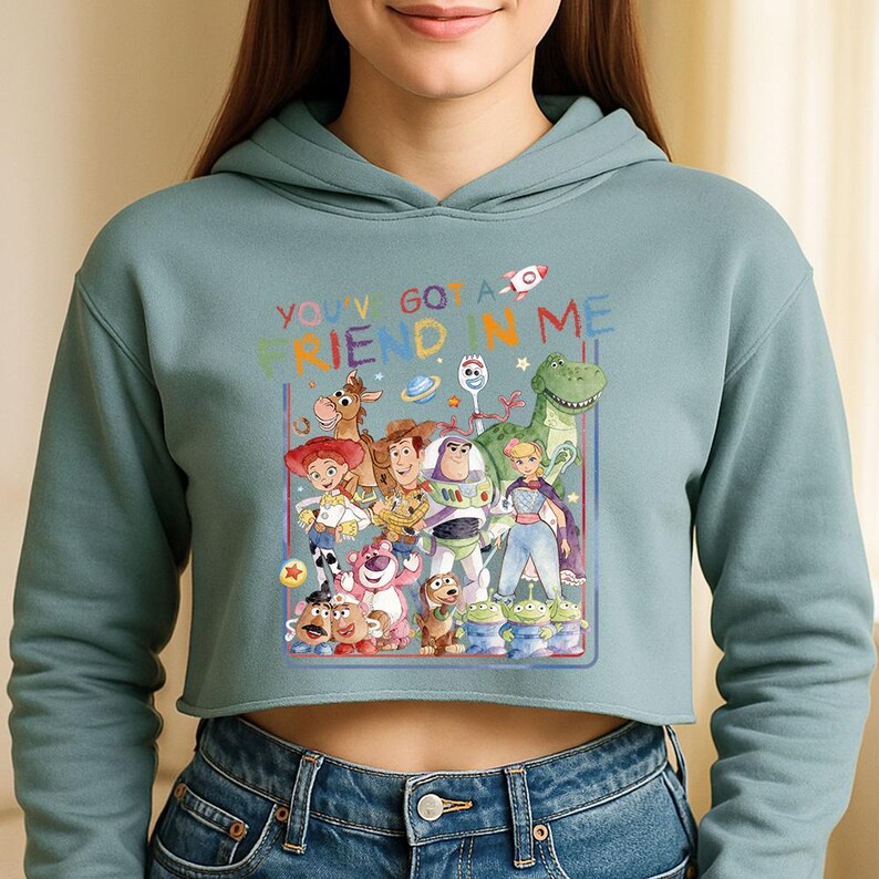 Może przedstawiać: Kr&oacute;tka, jasnoniebieska bluza z kapturem z kolorową grafiką postaci z filmu Toy Story. Grafika zawiera napis "YOU'VE GOT A FRIEND IN ME" oraz postacie z kresk&oacute;wek, takie jak Woody i Buzz Lightyear.
