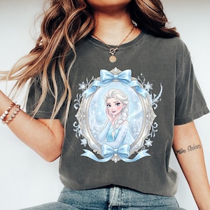 May include: Dark gray t-shirt featuring a portrait of a princess in a blue dress, framed by a light blue ribbon and ornate details. The princess has blonde hair and is smiling. The shirt is a comfortable, casual style.