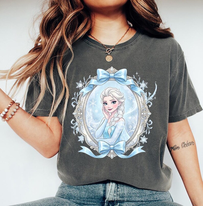 May include: Dark gray t-shirt featuring a portrait of a princess in a blue dress, framed by a light blue ribbon and ornate details. The princess has blonde hair and is smiling. The shirt is a comfortable, casual style.