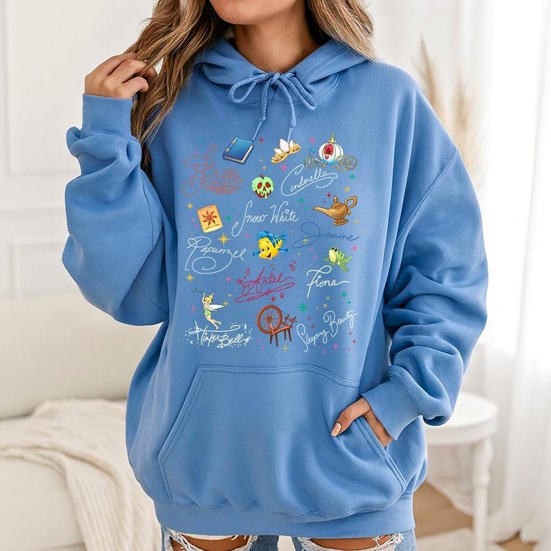 May include: A light blue hooded sweatshirt featuring a front graphic print of various animated character icons, including a golden lamp, a spinning wheel, a green apple, and a fairy, alongside cursive text names like Cinderella and Ariel.