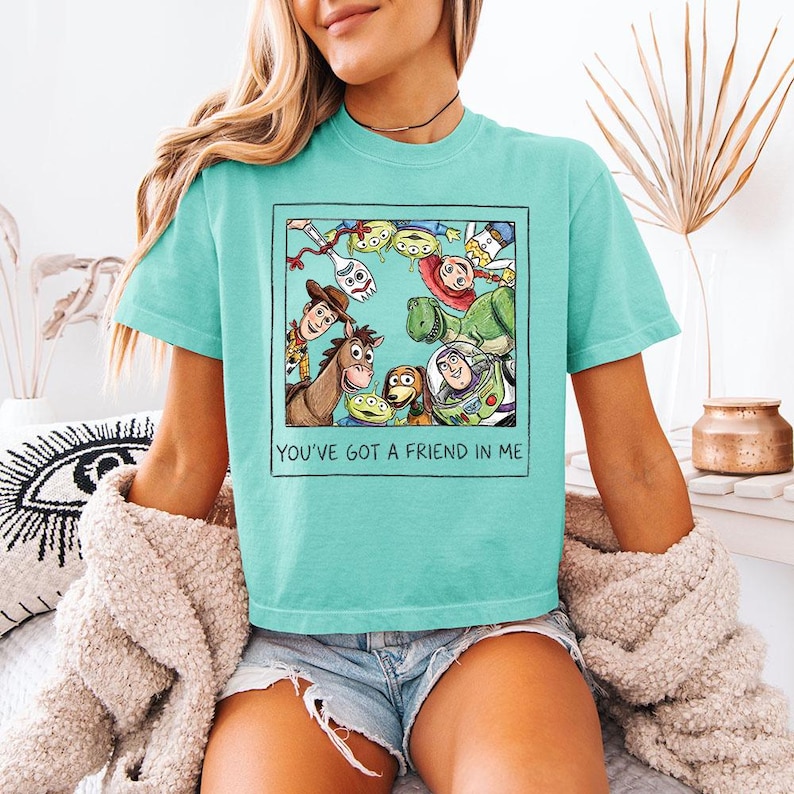 May include: A turquoise t-shirt with a graphic of characters from the animated film "Toy Story." The image includes Woody, Buzz Lightyear, and other characters, with the text "YOU'VE GOT A FRIEND IN ME" below the graphic.